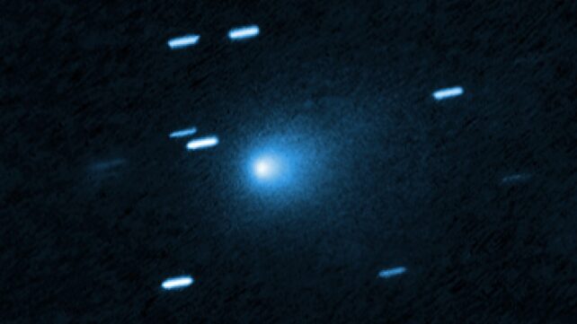 At the center of the image is a comet that appears as a teardrop-shaped bluish cocoon of dust coming off the comet’s solid, icy nucleus and seen against a black background. The comet appears to be heading to the bottom left corner of the image. About a dozen short, light blue diagonal streaks are seen scattered across the image, which are from background stars that appeared to move during the exposure because the telescope was tracking the moving comet.