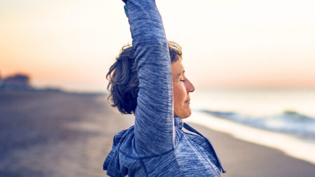 Woman stretching arms into air on a beach at sunrise. To illustrate news that scientists found a molecule that simulates exercise benefits for slowing aging