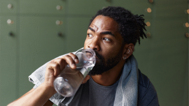 Black man with towel around his neck drinking from clear plastic water bottle in locker room with green lockers.