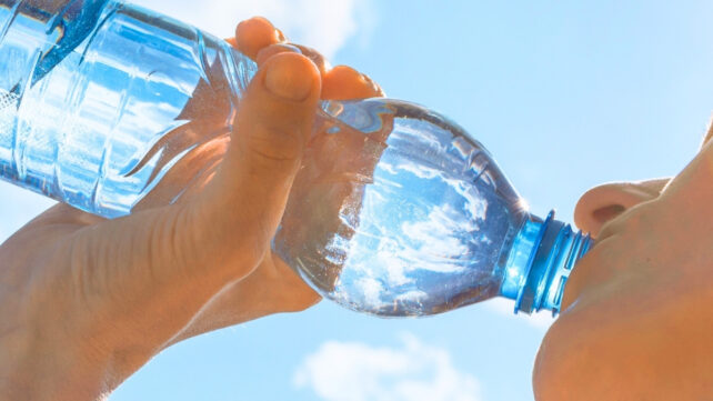 Hand holding a large bottle of water to mouth, with bright sky in the background