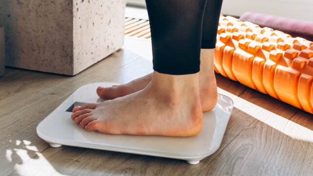 Feet standing on a weighing scale with dumbbells and fitness equipment in the background