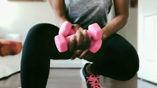 A woman crouched, holding two bright pink weights in one hand.