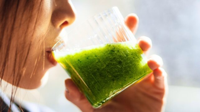 Woman drinking a green smoothie from a glass, against a high-contrast sunlit background