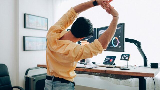 Man in yellow shirt at work desk, stretching arms overhead
