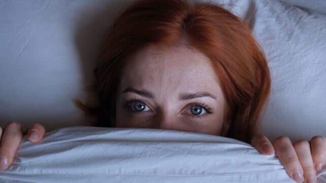 Sleepless woman with red hair lying in bed hiding under duvet at night