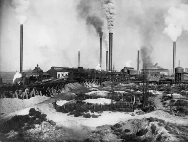 a black and white photo of an industrial smelting plant, with chimneys releasing smoke into the air.