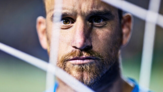 Determined adult male football goalkeeper looks through goal net directly at camera with stern and focused facial expression