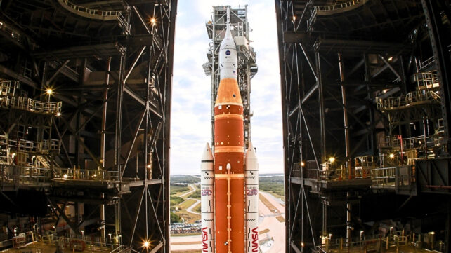 NASA's Artemis II rocket is brightly litin daylight in the center of the image, with high contrast equipment in the darkness of a building surrounding it