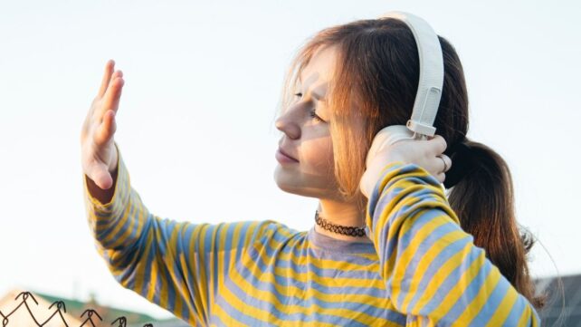 a white teen girl with brown hair in a ponytail is wearing over-ear headphones and holding her hand up, looking at the back of it. she is outside, in front of some solar panels and a chain link fence.
