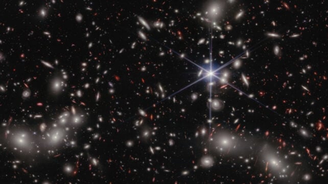 A crowded galaxy field on a black background, with one large star dominating the image just right of center. Three areas are concentrated with larger white hazy blobs on the left, lower right, and upper right above the single star. Scattered between these areas are many smaller sources of light; some also have a hazy white glow, while many other are red or orange.