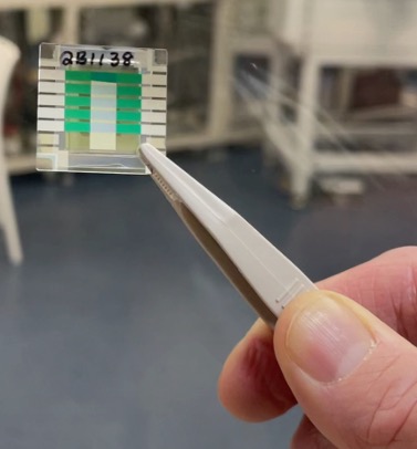 Close up image of hand using metal tweezers to hold a thin, square, green and white object.