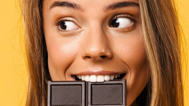 A young blonde woman biting down on dark chocolate