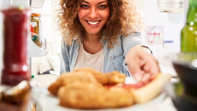View looking out from inside a fridge, of a young woman reaching out to grab leftover pizza