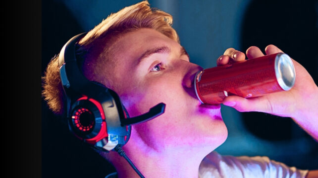 Young man drinking caffeine energy drink in high contrast purple light, helping him focus while gaming at home using headphones