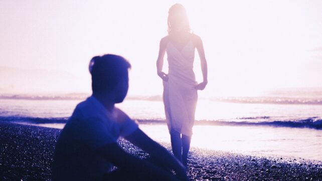 Silhouette of a man sitting on a seashore while looking at a woman in white dress walking towards him. The image looks like it could be capturing a distant memory