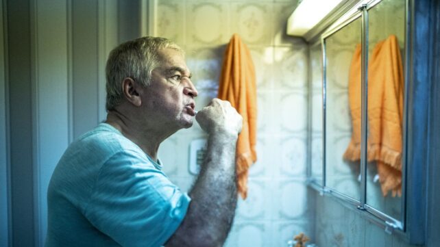 Grey-haired man brushing teeth in front of bathroom mirror, in low lighting and with orange towel hanging on wall.