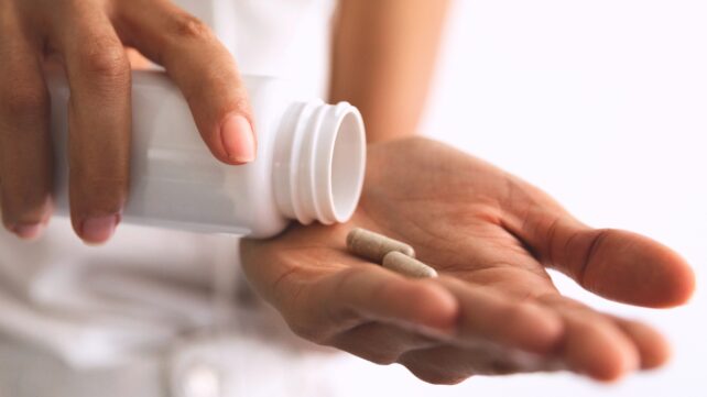 Hand with fingernails painted pink, holding a white pill bottle and tipping two supplement capsules into palm