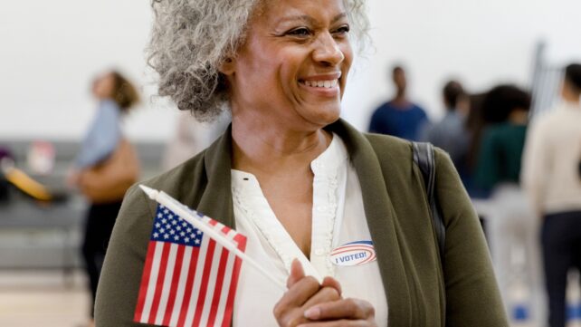 Older woman smiles while holding a US flag and wearing an “I voted” sticker at a polling place.
