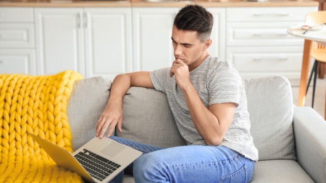 A young brunette man sitting on a gray couch, looking at an open laptop 
