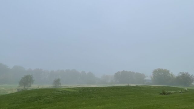 Fog blankets a green field and distant trees in a rural landscape, with a small building partly obscured in the background.