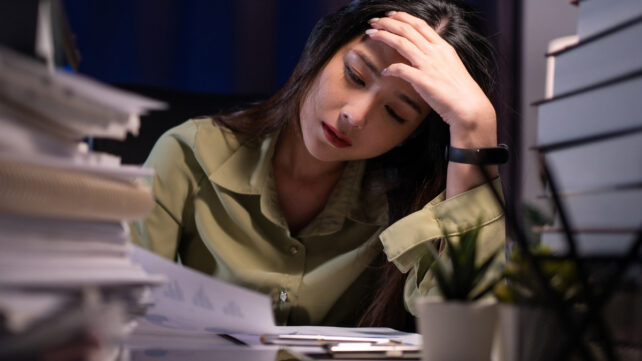 A woman in a green shirt looking tired, with a lot of work on her desk