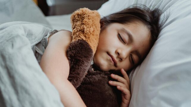 A young girl peacefully sleeping in bed with a stuffed teddy bear, showcasing tranquility and comfort.