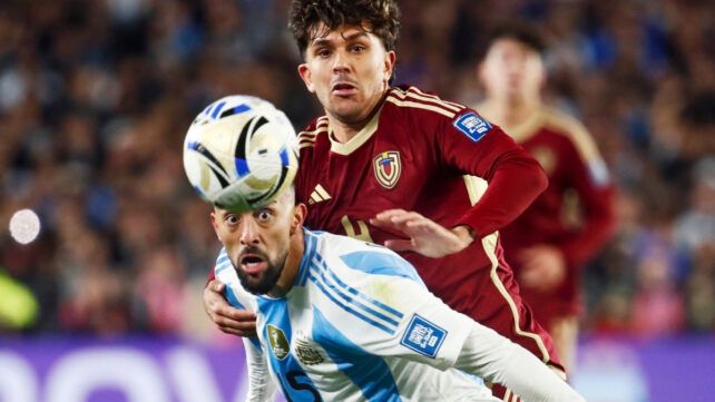 Two soccer players compete for the ball during a match, with one player in a light blue and white striped jersey leaning forward as another in a maroon jersey presses close behind him.