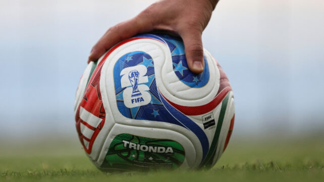 Close-up of a hand holding the official FIFA World Cup Trionda match ball on grass, with the red, blue and green patterned panels visible.