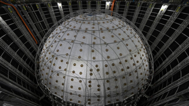 Large spherical JUNO neutrino detector inside an underground chamber, surrounded by cables, metal supports, and dark wall panels.