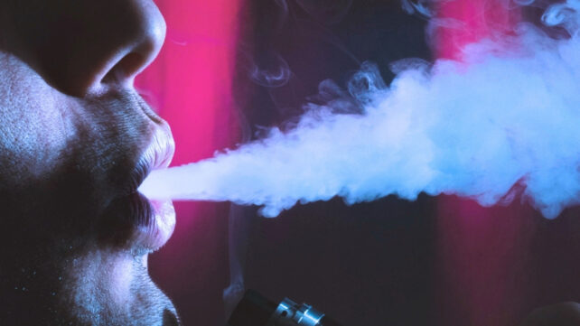 Close-up of a person exhaling a thick stream of blue-white vapor from a vape device against a dark background with pink lighting.