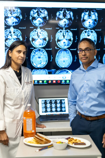 Sonia Anand and Russell de Souza standing behind a table of foods and an orange drink, with brain scans on a screen behind them.
